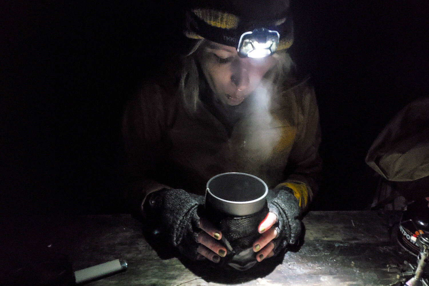 A hiker wearing the Petzl Tikkina Headlamp at night and blowing on a steaming cookpot