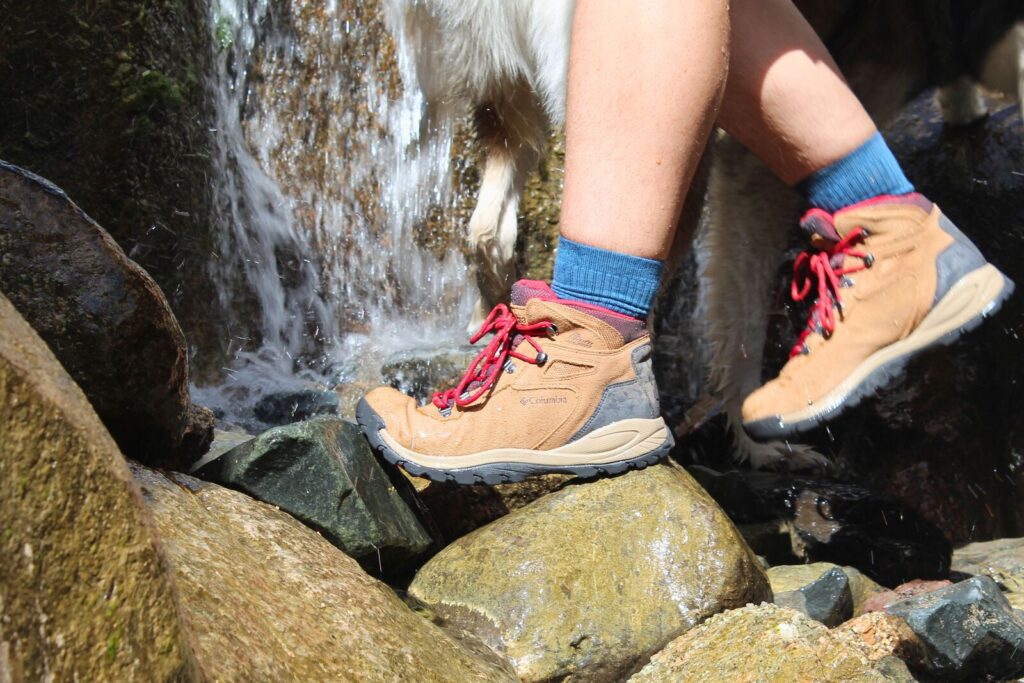 Hiker crossing over rocks in front of a waterfall wearing the Columbia Newton Ridge Plus Waterproof Amped