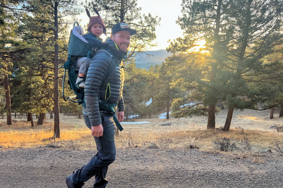 A scenic shot of a man carrying a child in a green Deuter Kid Comfort backpack carrier, with a golden sunset and trees in the background.