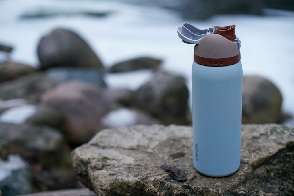 The Owala FreeSip Vacuum Insulated water bottle with its spout open sits on a rock with snow behind it.