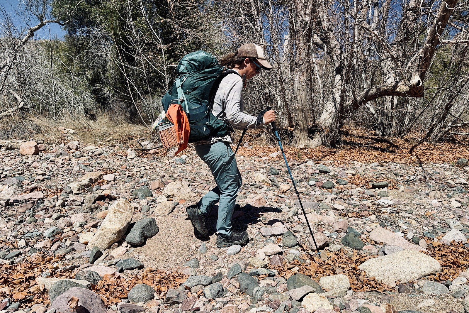 A person is walking in a rock filled wash wearing a backpack and using the Distance Carbon trekking poles. There are some trees in the background.