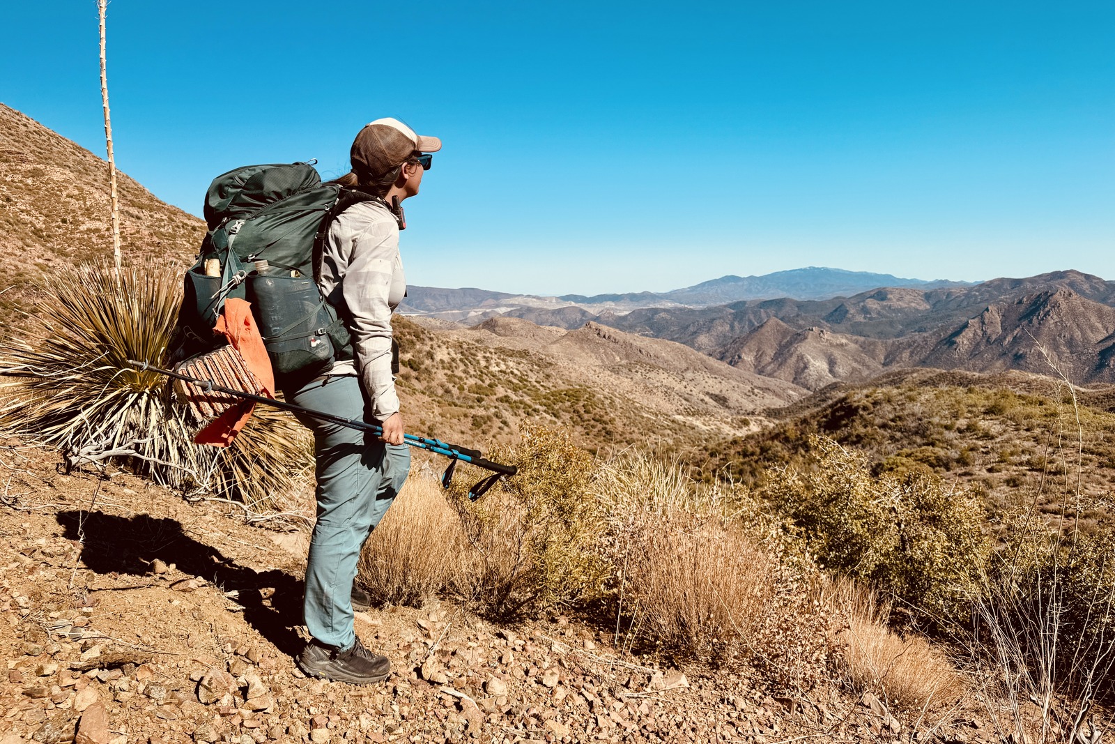 A hiker is standing looking out at the view while holding the Distance Carbon trekking poles. The area is desert like with mountains in the distance.