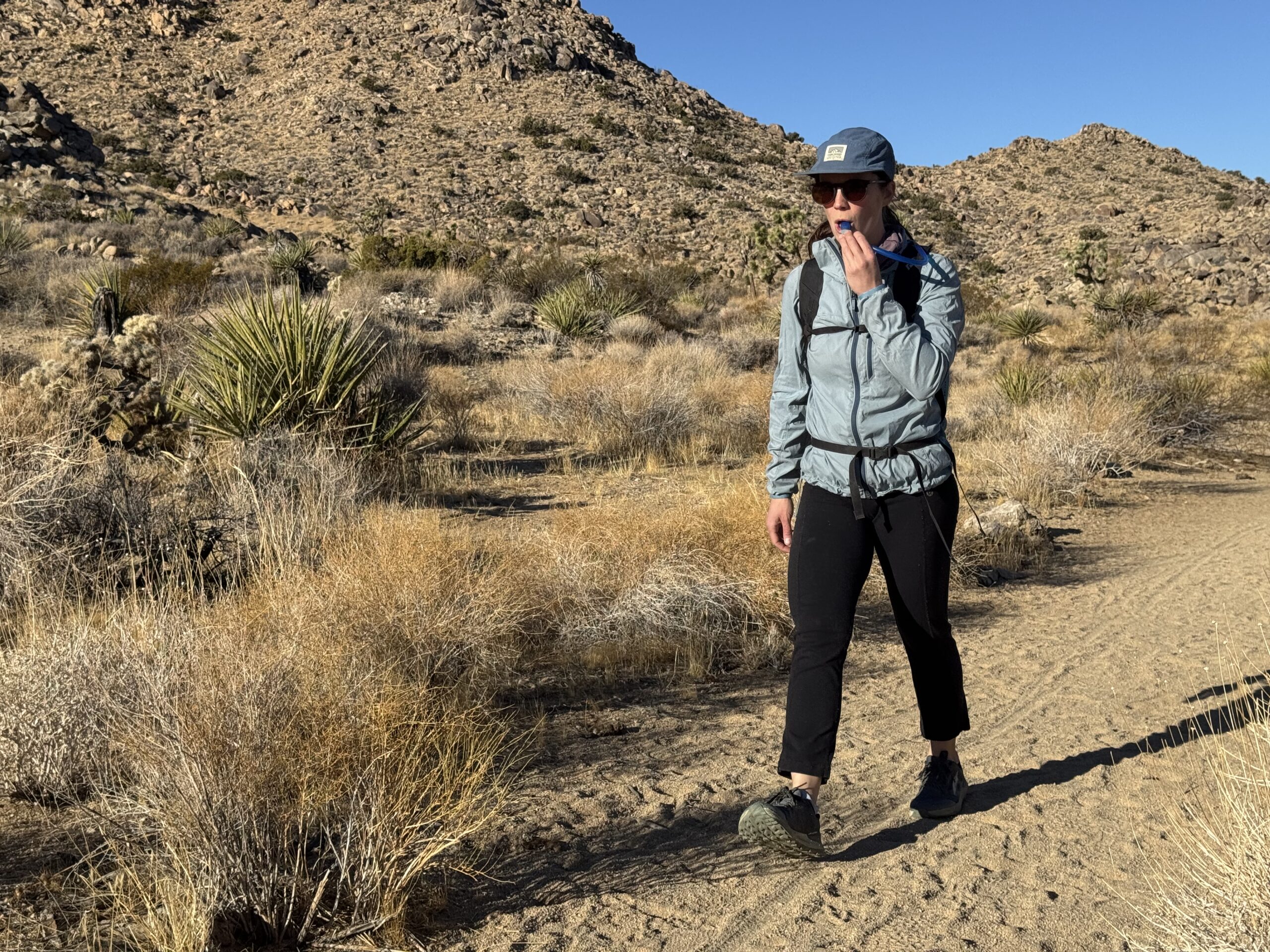 A hiker walks down a sandy trail in the Southern California desert while drinking from a hydration hose.