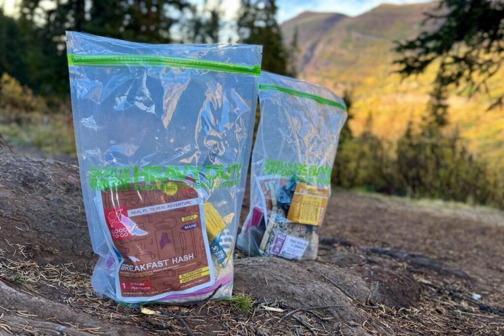 two smellyproof bags filled with food sit on the ground with mountains in the background