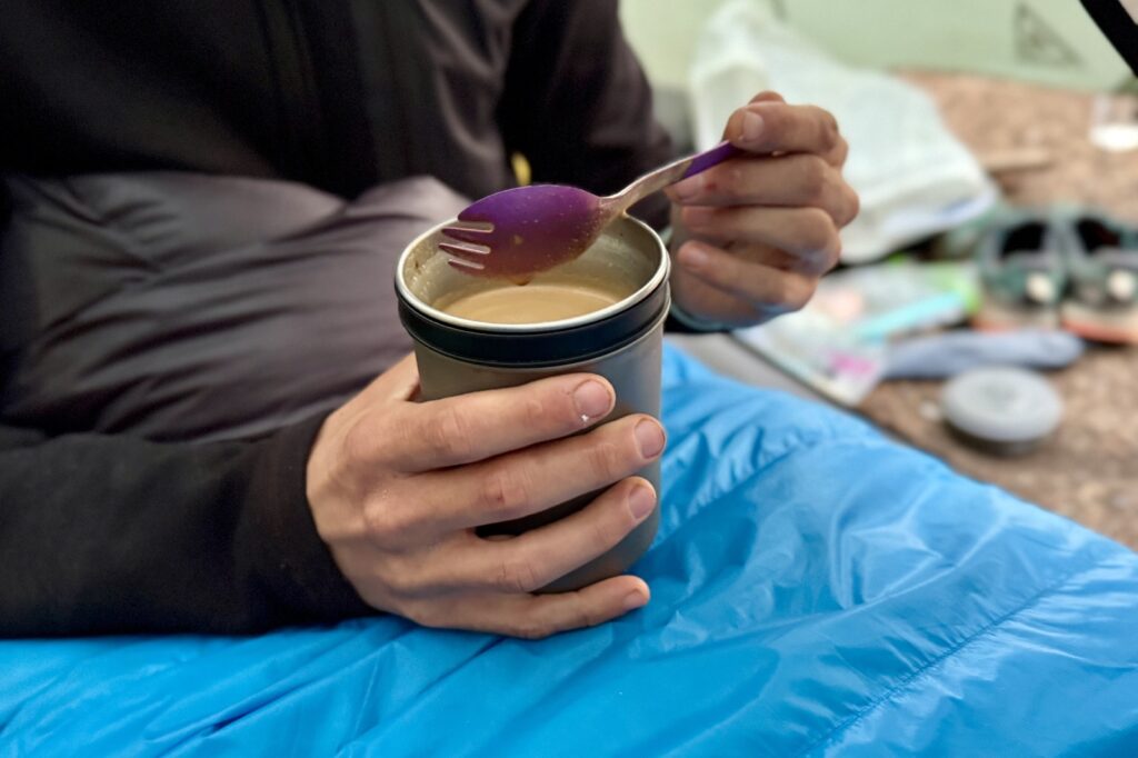 a chest down view of a hiker siting in a sleeping bag and stirring his coffee with a purple spork