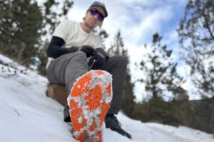 a hiker sits on a rock and shoes the bottom of his shoe with an ice cleat attached