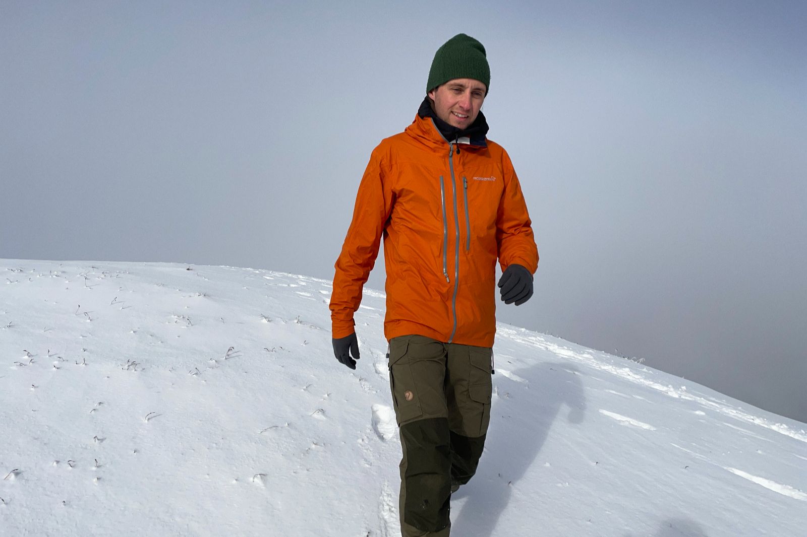 A man hikes down a hill through a snowy landscape wearing an orange jacket.