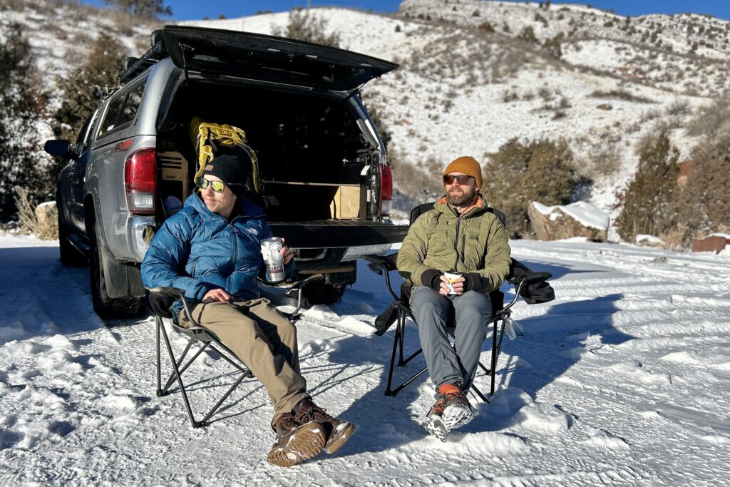 Two men sit in camping chairs at the back of a truck sipping drinks.