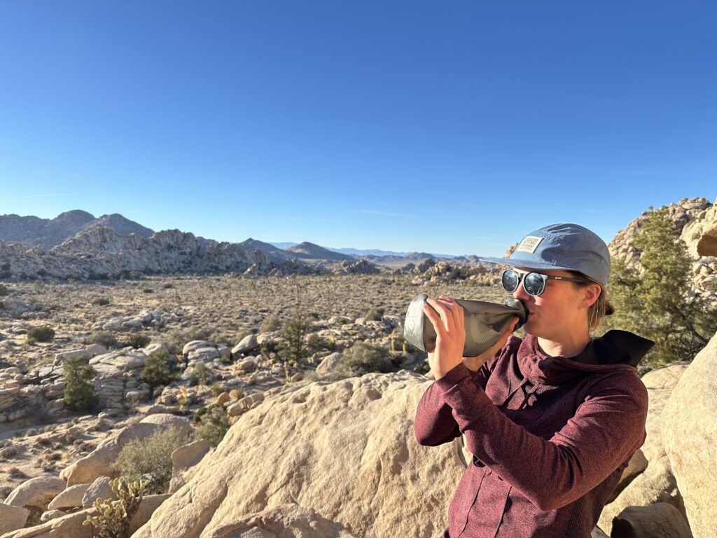 A person wearing sunglasses and a sun hat drinks from the HydraPak seeker water bag. The background is of Joshua Tree National Park, and it shows granite rock formations and some trees.