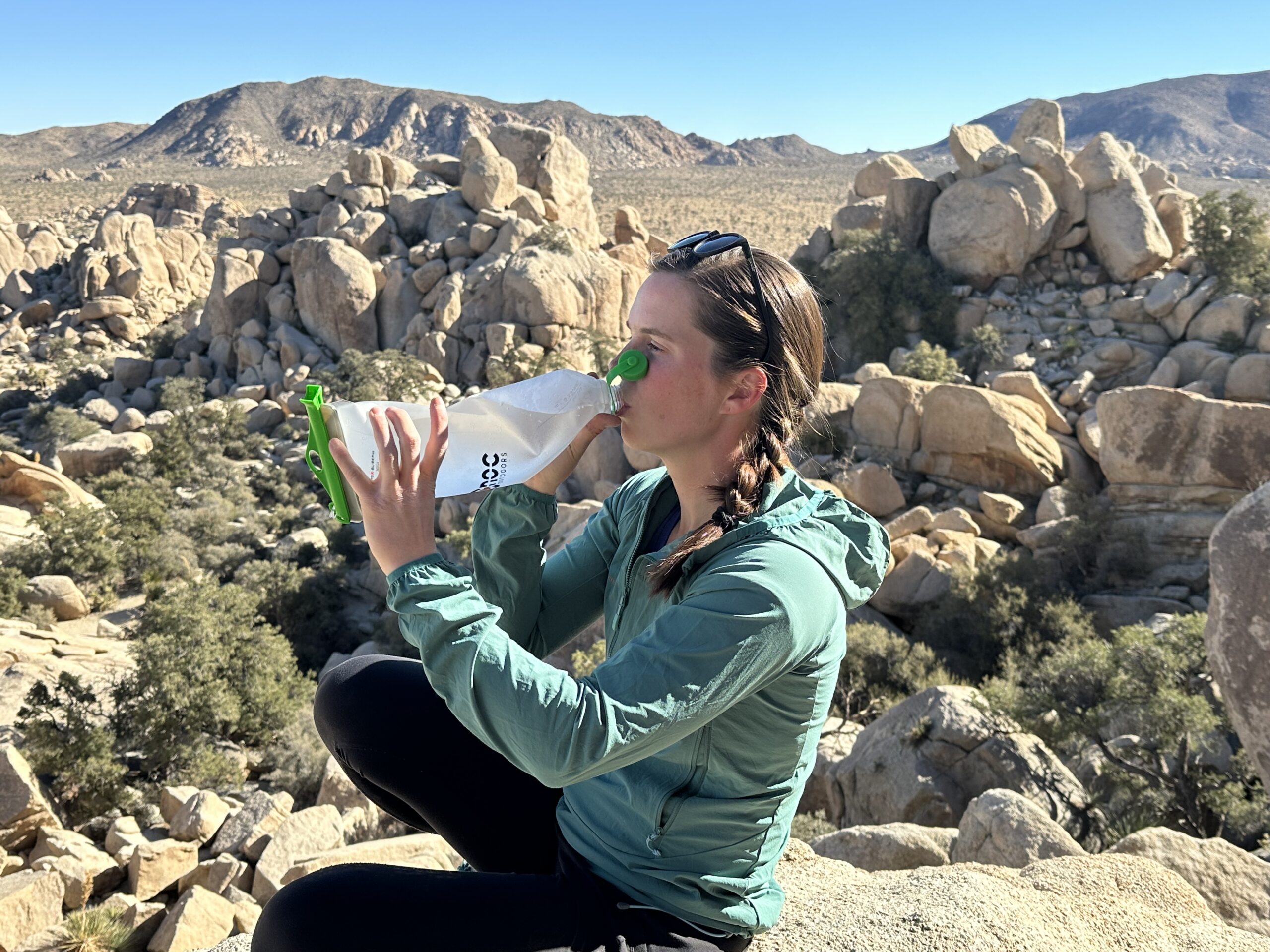 A hiker drinks from the CNOC VectoX water container, using two hands to hold it. The granite rock formations of Joshua Tree National Park are in the background.