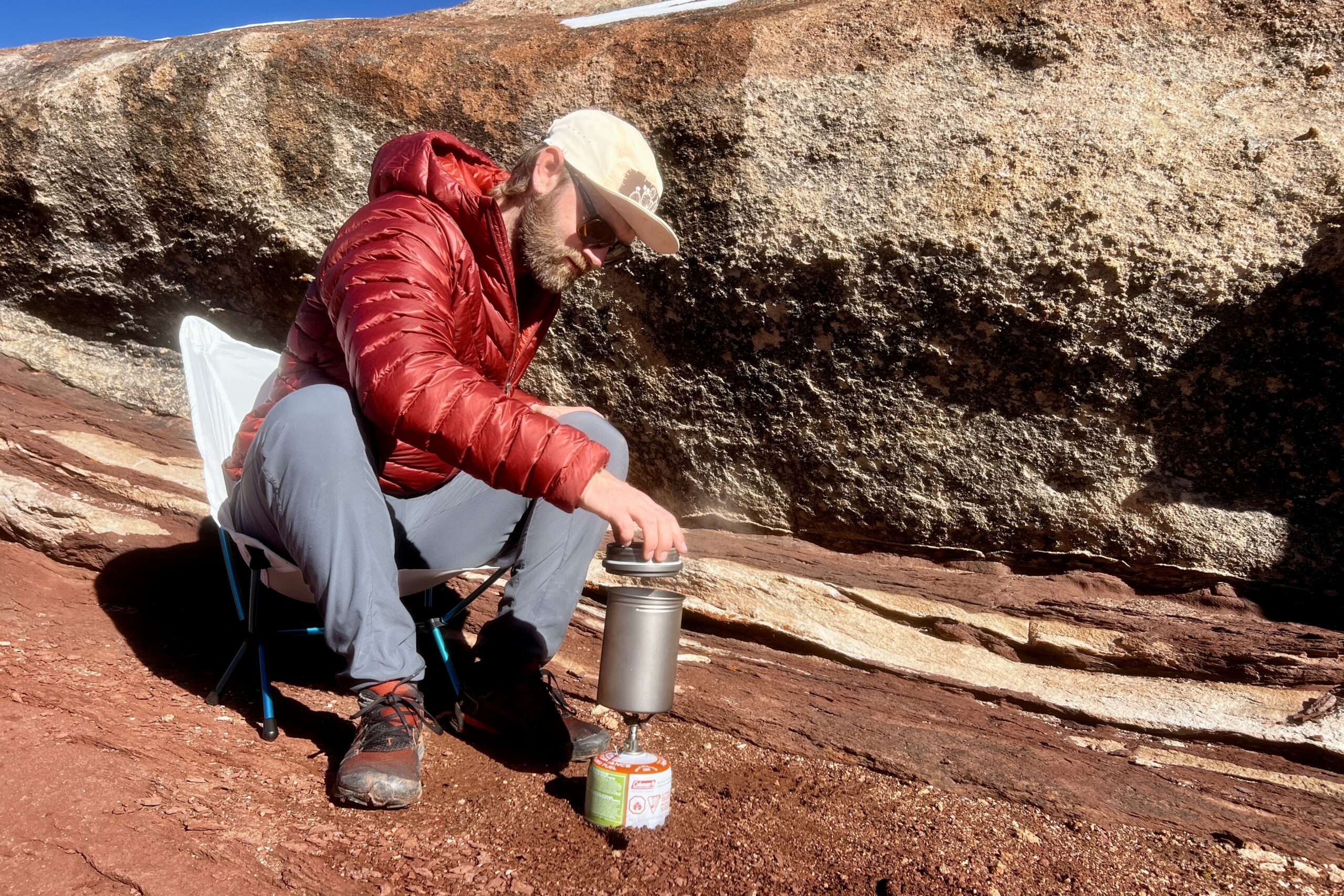 A hiker sits in a chair and pulls the lid off a backpacking pot against a red rocks background.