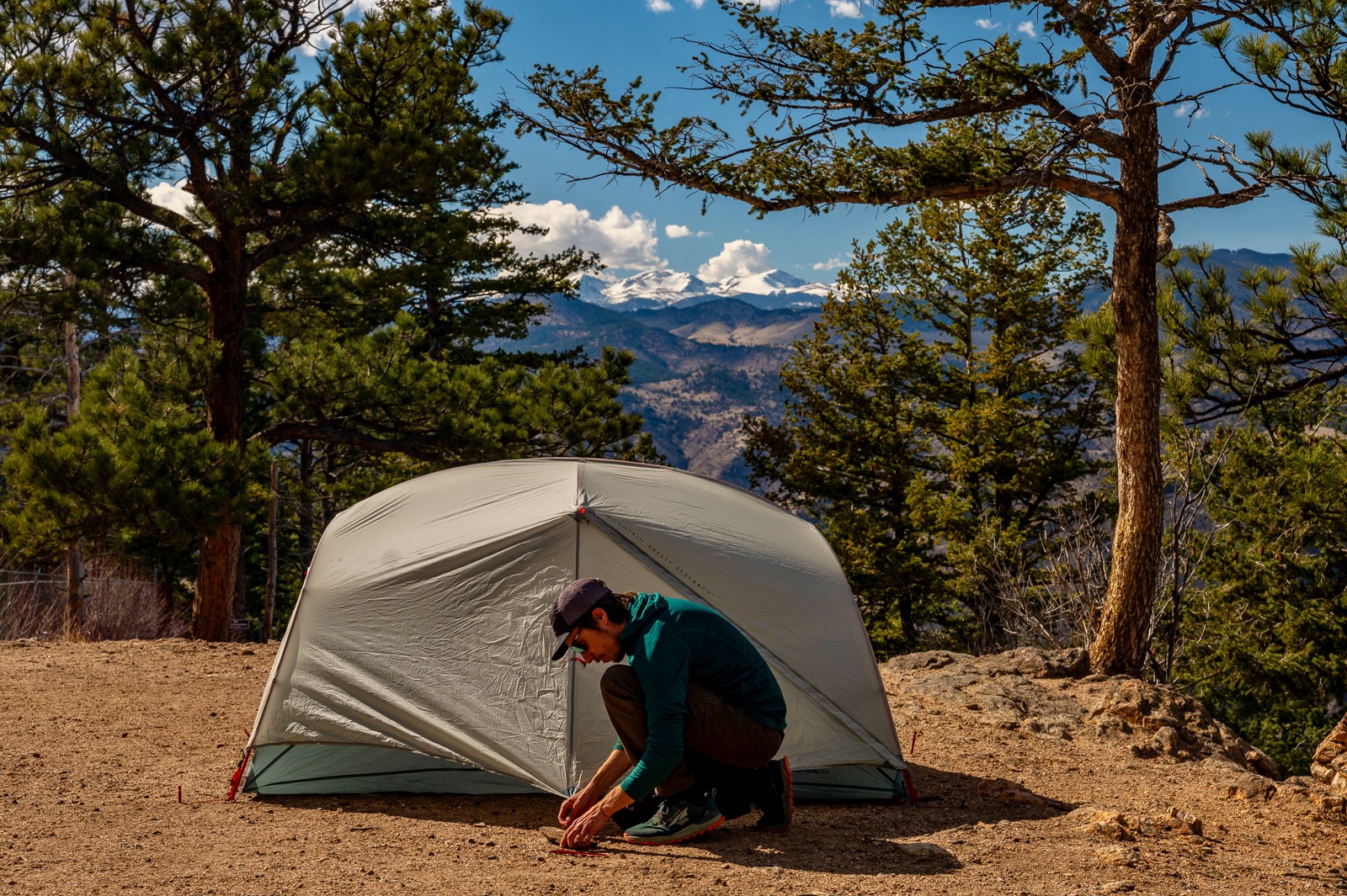 A hiker kneels down to stake out the vestibule of an msr tent with mountains and trees in the background.