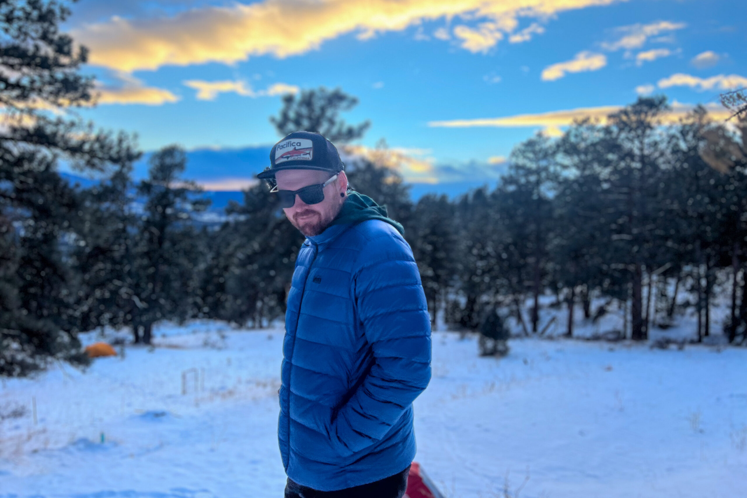 Person standing sideways in a snowy outdoor landscape wearing the REI 650 Down Jacket, hands in pockets, with sunset-lit mountains and trees in the background.